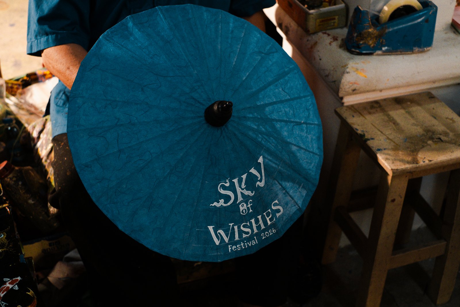 A deep blue paper umbrella, hand-painted with Sky of Wishes lettering, seen from above in the workshop